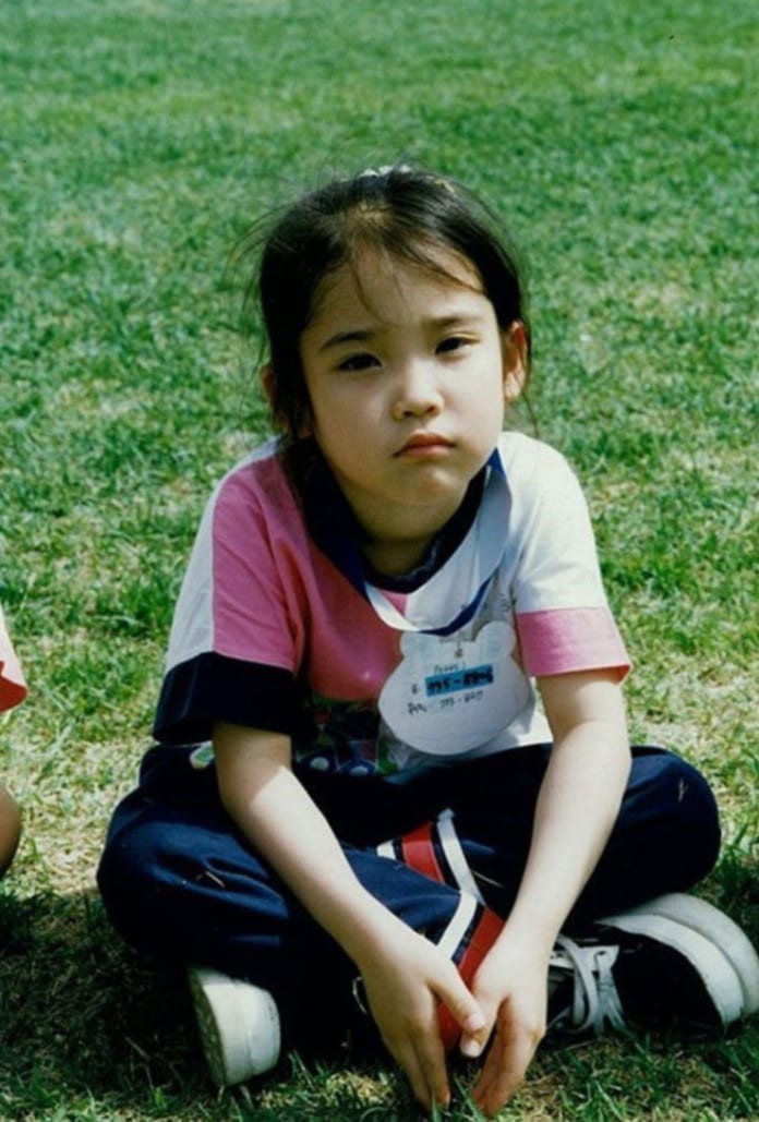 Ca sĩ IU hồi nhỏ, mặc quần áo thể thao ngồi trên bãi cỏ.