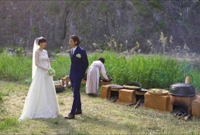 Won Bin và Lee Na Young mặc đồ cưới, đang nói chuyện cạnh bờ ruộng, phía xa là những bếp bằng đất nung nấu đồ cưới.