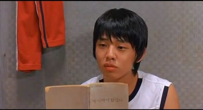 Yoo Ah In thời học sinh với mái tóc ngố, áo thun trắng và đang cầm cuốn sách.