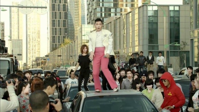 Ca sĩ Psy mặc quần hồng và áo khoác trắng, đứng trên nóc xe ô tô trong một MV của anh.
